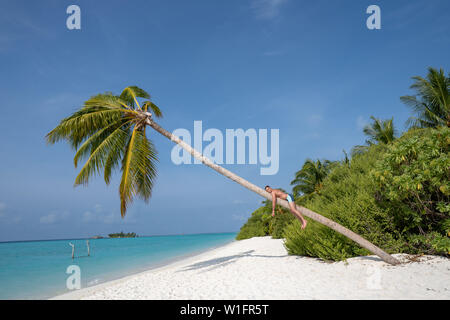 Un giovane ragazzo è appoggiata su di un grande albero di palma su una spiaggia di sabbia. In fondo è acqua turchese. Foto Stock