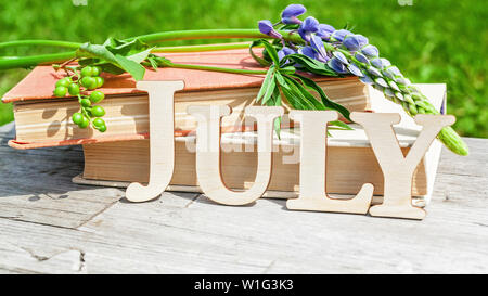 Parola luglio, una pila di libri vecchi e bouquet di fiori su un di legno bianco sfondo rustico. Composizione di estate Foto Stock