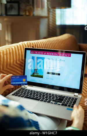 Donna con carta di credito utilizzando online travel hotel prenotazione sito su un computer portatile mentre è seduto sul divano del soggiorno moderno nella soleggiata caldo giorno d'estate. Foto Stock