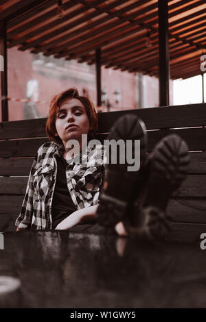 Giovane donna con corti capelli rossi in un plaid shirt in un bar di mettere le gambe sul tavolo Foto Stock