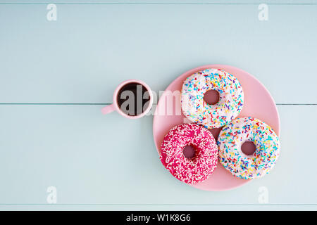 Ciambelle colorate in una piastra rosa rosa con cappuccino tazza da caffè su un pastello blu tavolo in legno sfondo con copia spazio. Vista aerea. Foto Stock