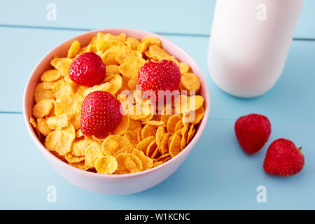 Fiocchi di mais cereale in una ciotola con topping di fragole e una bottiglia di latte sul blu del paese in legno stile tabella. Vista da sopra. Foto Stock