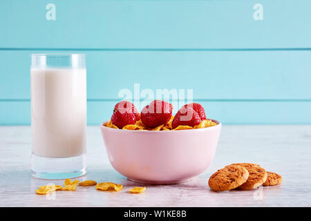 Ciotola di cereali secchi fiocchi di mais con topping di fragole, biscotti al cioccolato e un bicchiere pieno di latte sulla cucina in legno tavolo. Foto Stock
