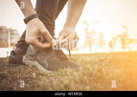 Un giovane uomo vestiti di nero è la legatura i lacci sul sneakers vicino atleta fitness seduta sul campo sportivo sull'erba. riscaldare corpo prepa Foto Stock