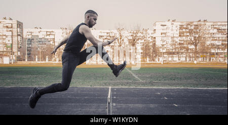 Un maschio caucasico in un salto oltre una barriera. in esecuzione sul Stadium. La via e il campo runner in sport uniforme in volo. energica attività fisica. Foto Stock