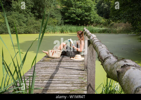 Giovane donna si rilassa sul molo di lakeside e legge Foto Stock