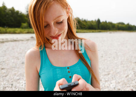 Giovane donna con un telefono cellulare al fiume Isar, Monaco di Baviera, Germania Foto Stock