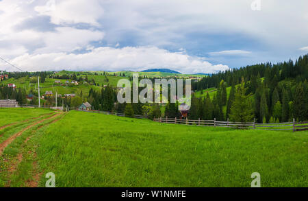 Molla di pittoresche montagne scena con split in legno cancellata attraverso una verde e lussureggiante pascolo, una strada di campagna e le vecchie case sulla valle susseguono Foto Stock