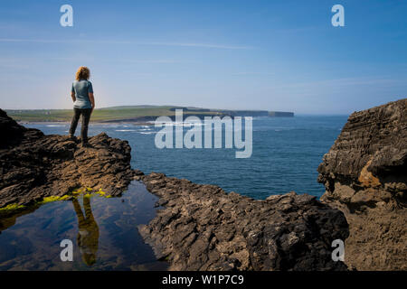 Al punto di vedetta vicino a Georges testa sulle scogliere di Kilkee, una donna sorge su rocce e si affaccia sul litorale, Byrnes Cove, Kilkee, County Foto Stock