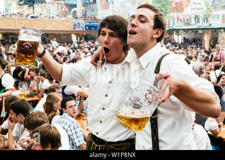 Giovani uomini in pantaloni di pelle in piedi sui banchi di birra Oktoberfest celebrare nella tenda della birra Foto Stock