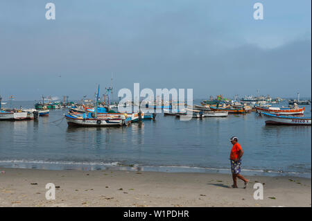 Un uomo cammina lungo la spiaggia di fronte a uno sfondo di barche da pesca, Paracas, Ica, Perù, Sud America Foto Stock