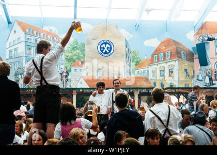 Giovani uomini in pantaloni di pelle in piedi sui banchi di birra Oktoberfest celebrare nella tenda della birra Foto Stock