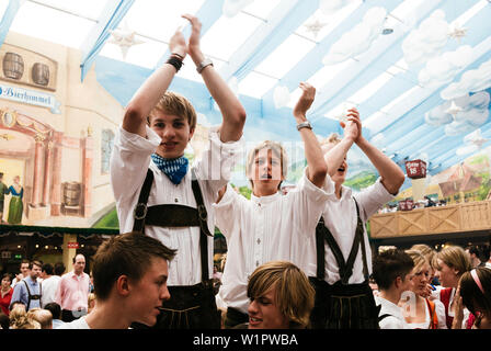 Giovani uomini in pantaloni di pelle in piedi sui banchi di birra Oktoberfest celebrare nella tenda della birra Foto Stock