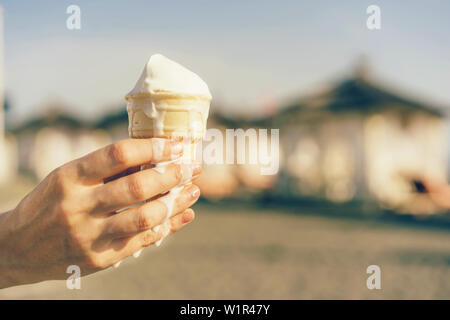 Cono di cialda con gelato in mano sullo sfondo della spiaggia tende, una calda giornata estiva, una massa di scongelamento fluisce lungo il braccio Foto Stock
