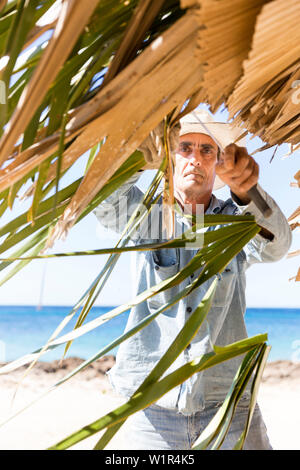 Uomo cubano di riparare il tetto di una pantina parasole realizzato da palme, solitario strada costiera di La Boca di Playa Ancon, con belle piccole spiagge di sabbia Foto Stock