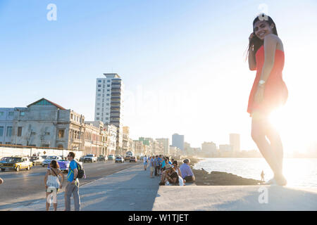 Giovane donna in un abito rosso che lo standig sulla parete malacon, turisti e gente locale e fisherman al Malecon, centro storico, centro città vecchia Habana Vieja Foto Stock