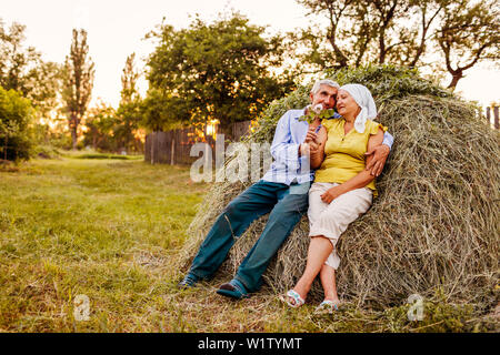 Coppia di famiglia di agricoltori seduti sul pagliaio e rilassante al tramonto in campagna. Duro lavoro di persone avvolgente. Agricoltura e concetto di agricoltura Foto Stock