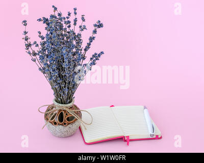 Fragrante bouquet di lavanda a secco con piccoli fiori viola in un bel vaso di ceramica bianco e una penna a sfera vicino al blocco di carta in un coperchio rosa Foto Stock