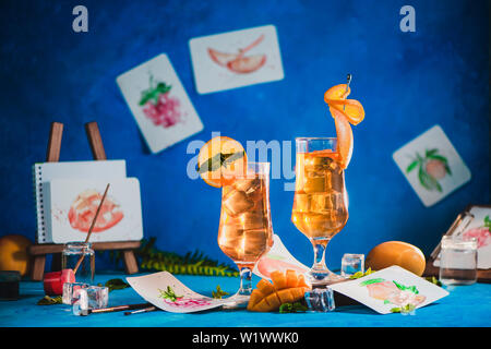 Estate bevande in bicchieri alti con schizzi di frutta in un artista workplace. mango e arancio tè freddo su uno sfondo blu con spazio di copia Foto Stock