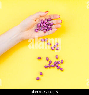 Donna mano azienda viola pillole manciata isolato in grassetto su sfondo giallo con copia spazio. Modello per la bellezza femminile blog social media. Hea femmina Foto Stock