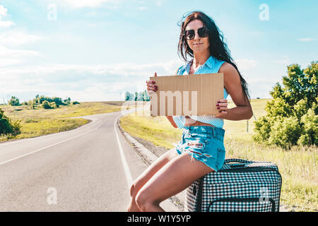 Piuttosto giovane donna autostop lungo una strada e in attesa su una strada di campagna con la sua valigia, cartone vuoto piastra. Bella ragazza in occhiali da sole il sollevatore Foto Stock