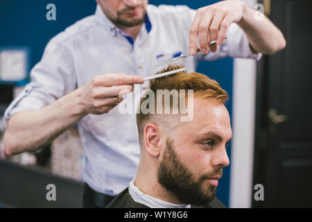 Barbiere rendendo taglio di capelli uomo barbuto in Barberia. Parrucchiere professionista il taglio di capelli di client in salone. Barbiere con le forbici e il pettine. Abile copricapo Foto Stock