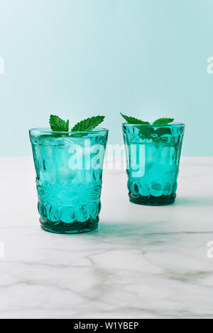 Due belle vintage occhiali turchese con bevanda fredda e cubetti di ghiaccio, decorata con fresco verde menta su marmo bianco tavola. Close-up. Foto Stock