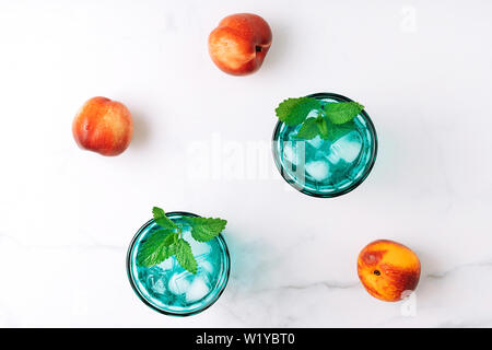 Due belle vintage occhiali turchese con bevanda fredda e cubetti di ghiaccio, decorata con fresco verde menta e tre pesche noci su marmo bianco tabl Foto Stock