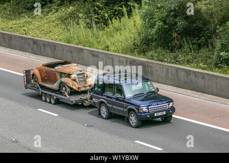 Rusty ha corroso il relitto di un'auto sportiva a due posti, automobili degli anni '1950 1930 Riley Sprite, un raro ritrovamento di un fienile, un progetto di restauro del veicolo, una vecchia auto d'epoca trainata da Land Rover d'epoca su un rimorchio per veicoli da ripristinare. Traffico veicolare nel Regno Unito, trasporto classico da collezione, condizioni di abbandono del collezionista, vecchie berline storiche sull'autostrada M6. Foto Stock