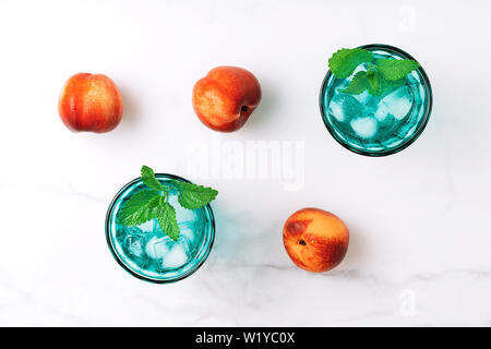 Due belle vintage occhiali turchese con bevanda fredda e cubetti di ghiaccio, decorata con fresco verde menta e tre pesche noci su marmo bianco tabl Foto Stock