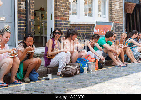 Londra Inghilterra - Luglio 15 2013; gruppo giovani adulti seduti lungo il marciapiede di rilassarsi e mangiare in città strada Foto Stock