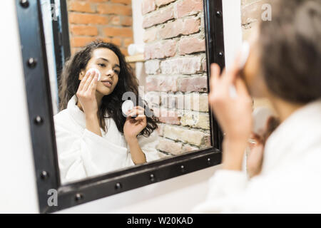 Bella giovane donna è la pulizia viso usando un dischetto di cotone e sorridere mentre guardando nello specchio del bagno Foto Stock