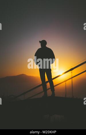 Silhouette di un giovane maschio in piedi sulla scala dietro scale con splendida vista del tramonto Foto Stock