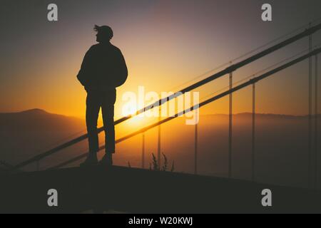 Silhouette di un giovane maschio in piedi sulla scala dietro scale con splendida vista del tramonto Foto Stock