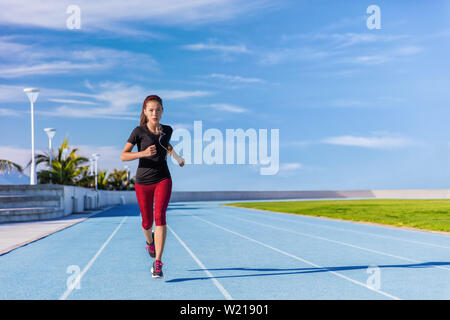 Femmina atleta asiatica runner in esecuzione su piste blu a stadio all'aperto in estate. Donna sportiva jogging ascoltando la musica con gli auricolari cardio training per la perdita di peso di successo. Il benessere e la salute. Foto Stock