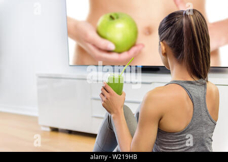 Mangiare sano donna bere frullato verde guardando la televisione online istruzione spettacolo TV su nutrizione e perdita di peso. Ragazza seduta a casa per imparare a perdere peso con frutta e verdura. Foto Stock