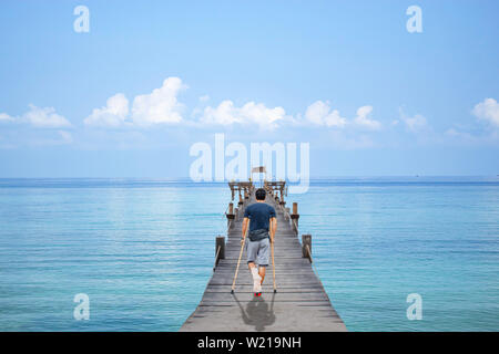 Uomo asiatico utilizzato stampelle in legno passeggiate sul molo ponte in barca il mare e il cielo luminoso a Koh Kood, Trat in Thailandia. Foto Stock