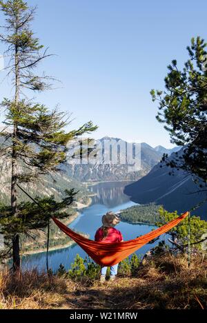 Donna con un cappello per il sole seduti in un arancione amaca, vista panoramica delle montagne con lago Plansee, Tirolo, Austria Foto Stock