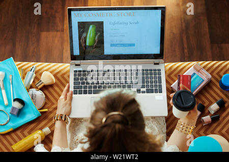 Vista superiore della giovane massaia in abito bianco con tazza di caffè rendendo il trucco di bellezza shopping online comodamente seduti sul divano nel soggiorno moderno. Foto Stock