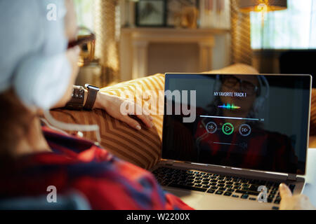 Sulla spalla shot del trendy hipster in rosso a scacchi shirt ascoltando la musica con le cuffie su un computer portatile mentre si posa sul lettino nel moderno liv Foto Stock