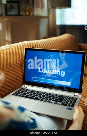 Triste casalinga moderna seduti sul divano di casa moderna nella soleggiata caldo giorno d'estate guardando al computer nella schermata di caricamento su un laptop. Foto Stock
