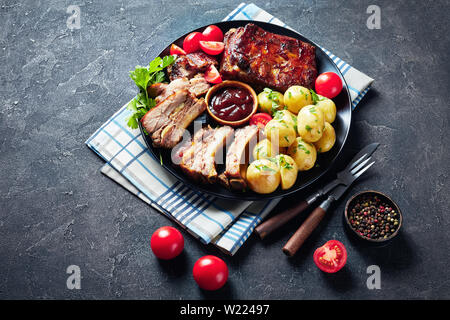 Deliziosa succosa carne di maiale alla griglia nervature condito con salsa barbecue serviti con pomodori e bollito di Hot Potatoes è servita su un piatto di nero su un calcestruzzo tabl Foto Stock