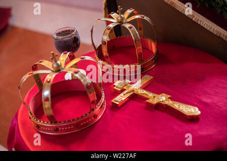 Croce, due corone e bicchiere di vino rosso sul tavolo preparato per il matrimonio. Chiesa ortodossa nozze accessori. Foto Stock