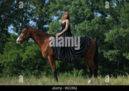 Giovane bella ragazza in abito nero giro sul cavallo nella foresta verde Foto Stock