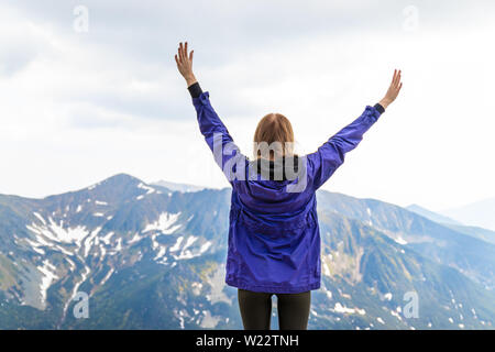 Giovane ragazza bionda travelerin una giacca blu ha sollevato le sue mani fino e godendo il verde paesaggio di montagna. Vista posteriore. Viaggi e turismo concept Foto Stock