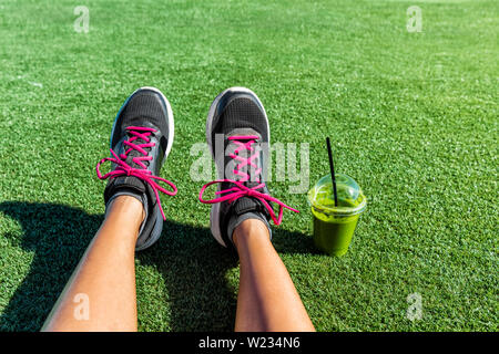 Uno stile di vita sano runner bambina scarpe running selfie con verde frullato. Donna Fitness di bere succo di frutta per andare dopo allenamento nel parco. Uno stile di vita sano sport atleta femminile pov telefono cellulare piedi foto. Foto Stock
