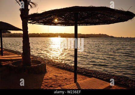 Un grande ombrello di vimini accanto ad un ponte di legno sedia sullo sfondo del mare e del sole al tramonto. Foto Stock