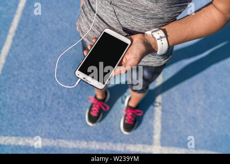 Runner ragazza tenendo lo smartphone utilizzando il touchscreen per la scelta della musica o i messaggi di testo SMS sulla prima app in esecuzione su pista. Atleta femminile donna piedi e gamba closeup con smartwatch e mano toccando il display. Foto Stock