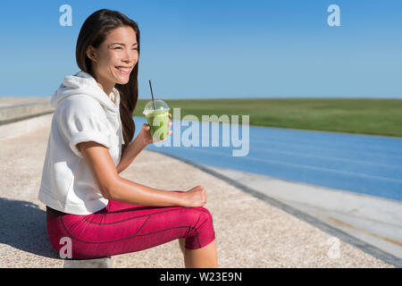 Una sana prima colazione al mattino runner atleta verde bere succo smoothie drink cup prima gara per un allenamento attivo. Esecuzione asiatica donna felice guardando stadium piste blu prima di allenamento cardio. Foto Stock