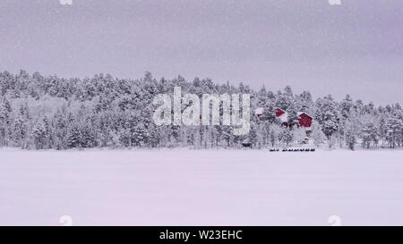 Finlandia, Inari - Gennaio 2019: vista attraverso il lago aperto di un dipinto di rosso di casa con una linea di motoslitte parcheggiate fuori come la neve continua a cadere Foto Stock